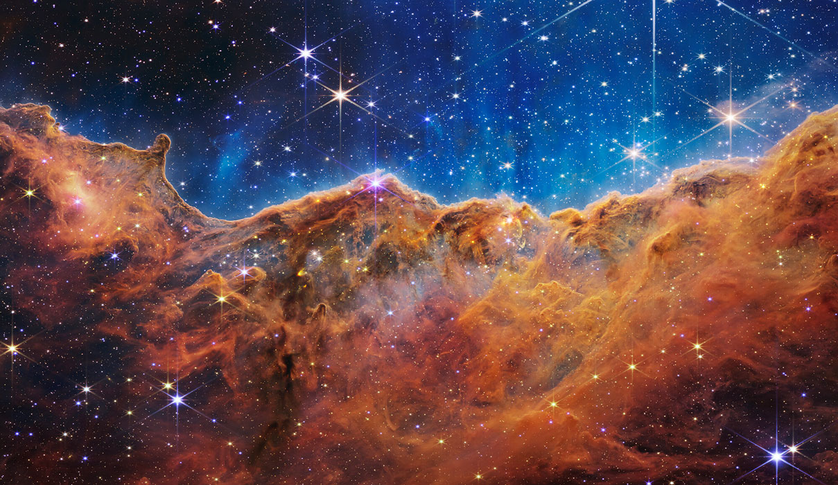 Ce paysage de “montagnes” et de “vallées” parsemées d’astres scintillants est le bord d'une jeune région de formation stellaire appelée NGC 3324, dans la nébuleuse de la Carène. Vue en infrarouge par le télescope spatial James Webb, cette image révèle pour la première fois des zones de naissance d'étoiles auparavant invisibles. Ces “Falaises cosmisques” sont le bord de la cavité gazeuse géante dans NGC 3324, et les "pics" les plus hauts de cette image mesurent environ 7 années-lumière de haut. La zone caverneuse a été creusée dans la nébuleuse par le rayonnement ultraviolet intense et les vents stellaires des jeunes étoiles extrêmement massives et chaudes situées au centre de la bulle, au-dessus de la zone montrée sur cette image.