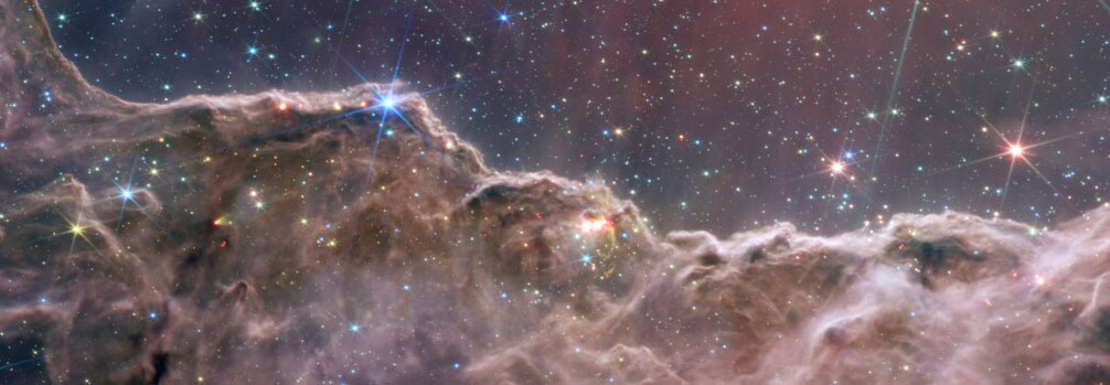 Une image composite des "falaises cosmiques" dans la nébuleuse de la Carène, créée avec les instruments NIRCam et MIRI du télescope Webb. Des nuages brun rosé de gaz et de poussière dominent le premier plan de l'image, scintillant de jeunes étoiles. Derrière, le ciel apparaît bleu marine, avec des étoiles brillantes et des galaxies. © Nasa/ESA/CSA/STScI